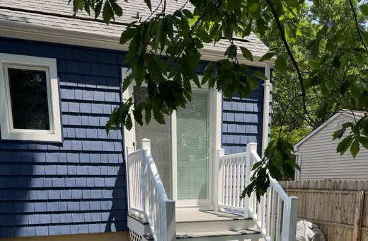 Newly built deck stairs with white vinyl railings and a paver landing at the base, on a blue shingle-sided home